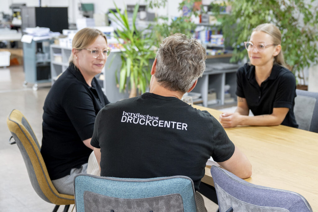 Zwei Mitarbeiterinnen und ein Mitarbeiter im Bergischen Druckcenter bei einer Teambesprechung am Tisch